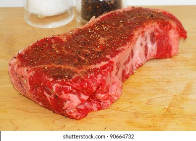 Large New York Strip Steak On Wooden Cutting Board With Salt, Pepper And Special Seasoning Being Added Prior To Cooking.