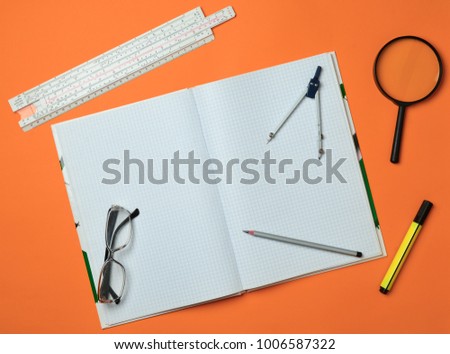 A large notebook for notes and drafting with stationery objects for a student on an orange background. Ruler, marker, pencil, magnifying glass, compasses, glasses. Top view.