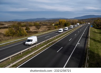 A large number of minivans move along the highway carrying goods for all people in the world. White modern delivery small shipment cargo courier van moving fast on motorway road to city urban suburb. 