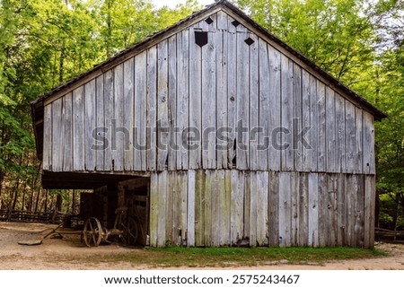 A large, old barn with a wheelbarrow in front of it. The barn is made of wood and has a rustic, weathered appearance. Concept of nostalgia and a connection to the past