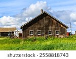 A large, old, wooden house with a chimney sits in a grassy field. The house is surrounded by a fence and a few other buildings. The sky is cloudy, and the grass is lush and green