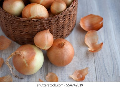 Large Onion Harvest In A Wicker Basket. Bulb Onion Is Rich In Vitamins, Useful Spring. Onion Peel On A Wooden Background. Large Onions Can Be Seen From Above.