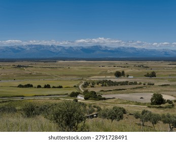 A large open area features fields and a small house located at the edge. Mountains rise in the background under a clear blue sky. A road runs through the landscape.