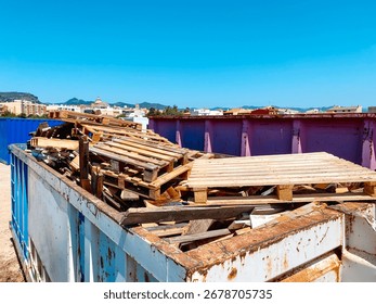 a large open container filled with broken wooden pallets and used wooden planks, located in an outdoor recycling center, with town buildings and mountains visible in the distance