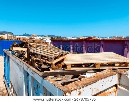 a large open container filled with broken wooden pallets and used wooden planks, located in an outdoor recycling center, with town buildings and mountains visible in the distance