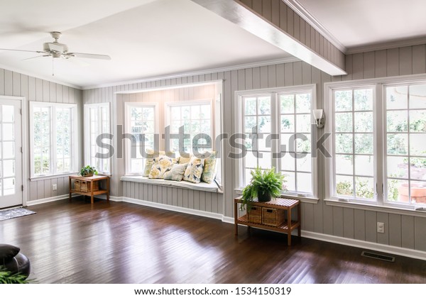 Large\
and open living room den sun room with windows on two sides and\
lots of natural light flowing in. There is a window seat on one\
side and a leather couch and plant on the\
other.