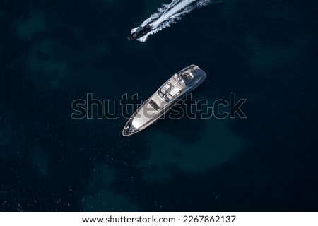 Large open yacht anchored in the blue sea top view. Silver modern boat on blue transparent water aerial view. Boat fast moving next to yacht top view.