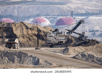 Large open-pit mining scene featuring a power shovel loading a dump truck amid spoil piles and storage domes; Mining in British Columbia, Canada with heavy equipment and industrial extraction activity.