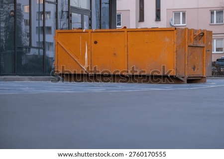 Large orange construction waste container placed near a residential building in urban area. Scene represents renovation, waste management, and city infrastructure. Suitable for construction, demolition, recycling, and urban development concepts. Ideal for use in industrial, environmental, and city planning related content. 