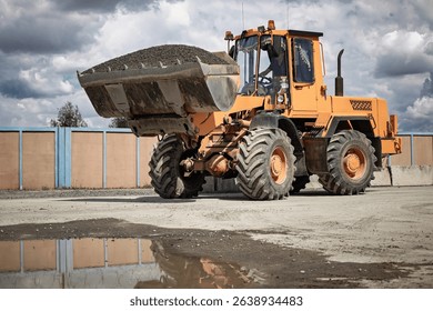 A large orange loader is moving gravel at a construction site, surrounded by a gray sky and reflective puddles on the ground.