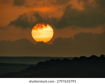 Large orange sun setting behind distant hills under dramatic sky