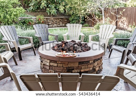 Large outdoor fire pit surrounded by wooden rocking chairs, beautifully landscaped backyard, with the glass of wine and food platter.