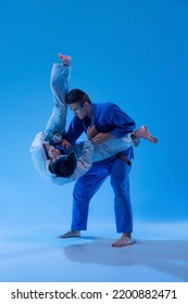 Large Outer Reap. Two Sportsmen Training, Practising Martial Arts, Judo Isolated Over Blue Studio Background In Neon Light. Concept Of Martial Art, Combat Sport, Health, Strength, Energy, Fit.