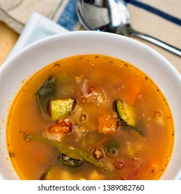 A Large Overhead View Of A Round White Bowl Of Minestrone Soup With Vegetables, Sliver Spoon, Blue And Tan Cloth Napkin.