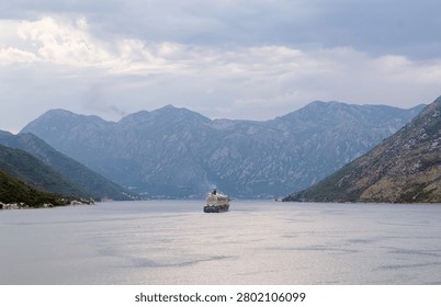 Large passenger liner navigating through the calm waters of Kotor Bay surrounded by massive mountains. Cruise ship in Montenegro under an overcast scenic sky