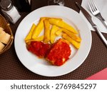 Large piece of cod fillet cooked with tomato sauce. Portion of fried French fries and glass of chilled beer are served with dish.