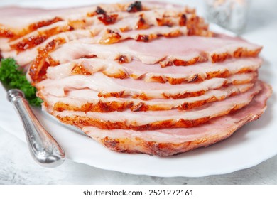 A large piece of ham is sliced into thin pieces and served on a white plate. The ham is glazed and has a crispy texture. The plate is set on a table with a silver fork and a sprig of parsley