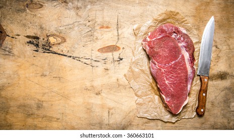 A Large Piece Of Raw Meat With A Butcher Knife On The Paper. On A Wooden Table. Free Space For Text . Top View