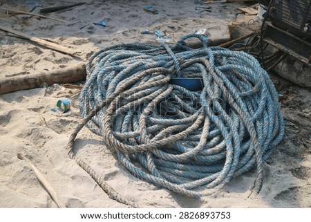 Large pile of coiled thick blue and grey nautical rope resting on a sandy beach near fishing machinery. Tangled heap of old marine ropes on the sand, ideal for concepts related to seafaring, maritime industry, and fishing gear.