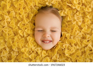 A large pile of dry noodles in the form of a bow. Pasta made from durum wheat. The child's face is surrounded by a large amount of pasta. The funny girl squeezed her eyes shut. Smile of the child
