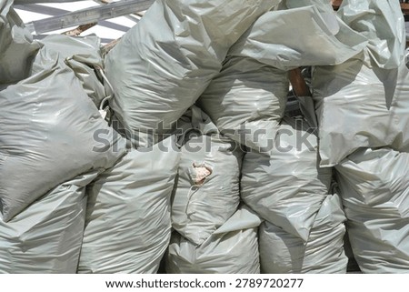 A large pile of filled gray plastic rubble sacks stacked together with one bag showing a small torn hole revealing construction waste