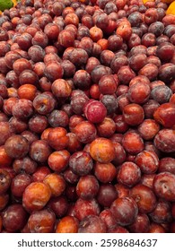 A large pile of fresh, ripe plums with a natural dusty bloom, highlighting their deep red and purple hues. The plums have a smooth yet slightly matte skin, indicating freshness and ripeness.