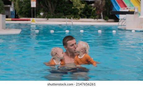 In A Large Pool With Clear Blue Water, The Father Keeps Two Twin Girls, Kisses The Girls In Turn And The Three Of Them Enjoy Swimming And Smile Sweetly. Happy Holidays With Kids.