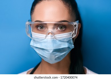 large portrait of a young girl, brunette with gathered hair in safety glasses with green eyes, a protective medical mask looks straight, dressed in a white long sleeves on a blue background soft focus