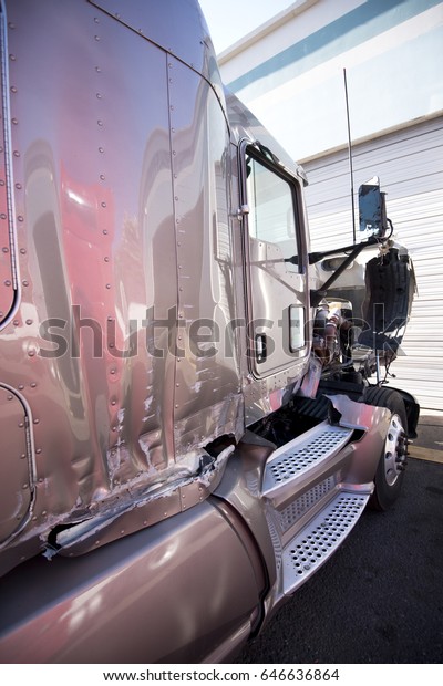 A large professional semi truck crashed in a car
accident stands with an open damaged hood and a crumpled body near
the garage gate