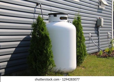 A Large Propane Tank On The Side Of A House Bordered By Two Cedar Bushes.  The House Has A Bluish Grey Siding