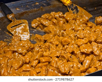 A large quantity of meat chunks being cooked in a thick, creamy orange-brown gravy sauce on a flat-top griddle.	
