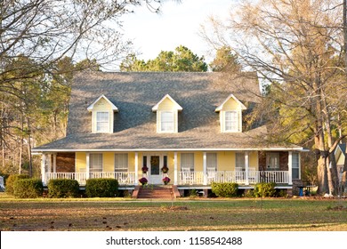 A Large Ranch Style Country Home With A Wrap Around Porch And Dormer Windows. The Large Yard Features A Mature Landscape With Trees, Bushes And Grass.