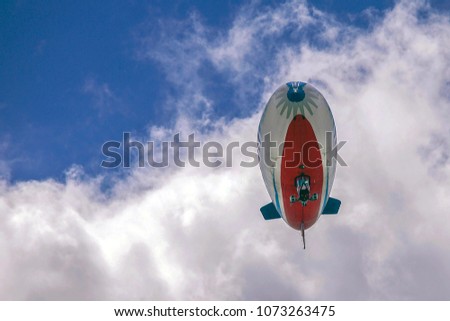 Large red and blue blimp flying amongst the fluffy white clouds in a blue summer sky.