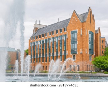 A large red brick building with tall windows and a slate roof stands behind a water fountain spraying jets on an overcast day.