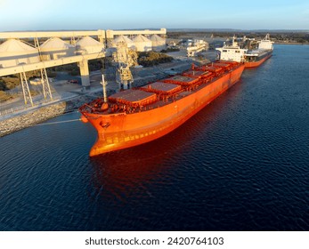 A large red bulk cargo ship docked at an industrial port facility, positioned beside a loading structure equipped with conveyor systems and tall storage silos used for transferring bulk materials. Sunlight highlights the ship’s hull, the machinery, and the surrounding industrial buildings, while another vessel sits moored behind it. The calm water and clear sky create a bright, detailed view of maritime transport infrastructure, showcasing the scale and complexity of global shipping and bulk material handling.
