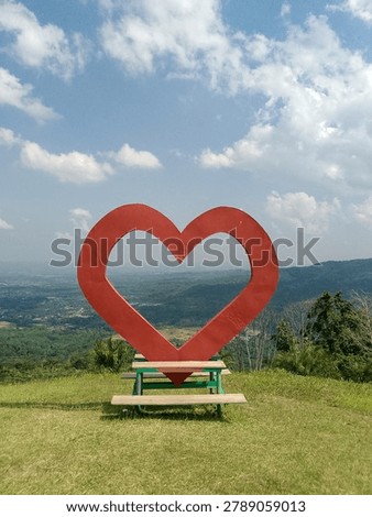 Large red heart-shaped frame overlooking a scenic mountain and valley landscape under blue sky.