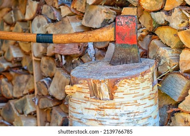 A Large Red Metal Axe With Some Rust On The Iron In The Center Of A Large Birch Trunk. There's A Stack Of Junked Wood Behind The Axe And Tree Stump. The Wooden Handle Has Black Tap On The Handle.