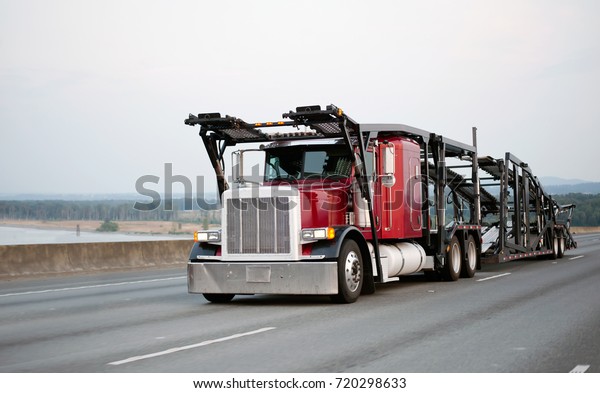 A large red powerful big rig car hauler\
semi truck with an empty two-tiered trailer for transporting cars\
is moving along the broadband scenic highway to the loading point\
of cars for transportation