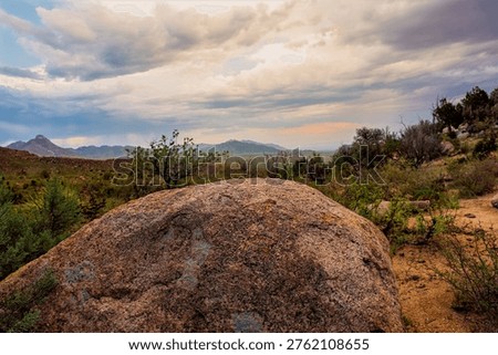 A large, reddish-brown boulder sits in the foreground of a desert landscape under a cloudy sky. The boulder is textured and appears to have some lichen or moss growing on it. In the background, rolling hills and mountains are visible, dotted with sparse vegetation and trees. The sky is filled with dramatic, heavy clouds, suggesting an approaching storm or the aftermath of one. The overall scene evokes a sense of rugged natural beauty and the vastness of the desert environment.