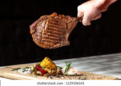 A Large Roasted Piece Of Mutton Steak, Pork, Veal Or Beef On Bones Cooked On An Air Fryer, In The Hands Of The Chef's Chief, A Background Image On A Black Background.