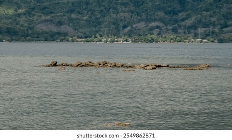 A large rock formation sits on the surface of a large body of water. The water is calm and the sky is overcast - Powered by Shutterstock