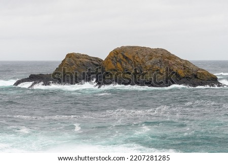 Large rocks in the teal colored ocean are covered in green moss and orange algae. The waves are white and rough hitting the small island. The ocean is stormy and the sky is grey and foggy.