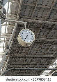 A large round wall clock hanging from the ceiling of a train station or industrial-style building. The image conveys a sense of punctuality, travel, and urban infrastructure.