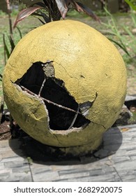 A large, round, yellow object, seemingly a decorative sphere made of concrete or plaster, is cracked and broken in several places.  Visible through the cracks is a metal reinforcement cage.