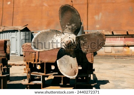 Large, rusted ship propeller displayed on wooden blocks in a shipping yard, symbolizing industrial maritime decommission.