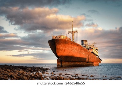 A large, rusted shipwreck rests partially submerged near a rocky shore under a dramatic, cloudy sky at sunset, casting a golden hue over the scene. - Powered by Shutterstock