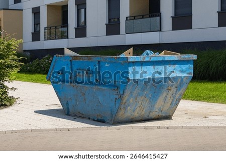 Large rusty blue dumpster container placed near modern apartment building on sunny day. Residential waste management, urban maintenance, cleanliness and outdoor property organization