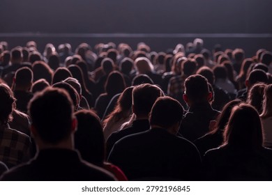 Large seated audience watching performance in dim auditorium, silhouettes and backlit heads create quiet, focused atmosphere.