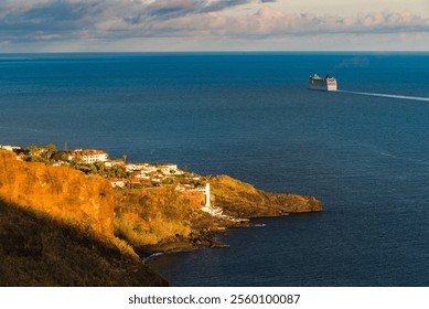 A large ship leaves the shores of Madeira, gliding through calm waters as the sun sets behind the rocky landscape and charming coastal village. The warm light enhances the scenery. - Powered by Shutterstock