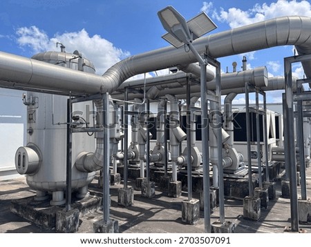 Large, silver industrial processing vessel and insulated pipes connect to rooftop HVAC units, supported by steel frames and concrete pillars. A solar-powered street light stands nearby under a bright 